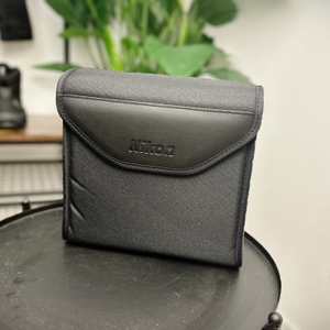 Die Nikon Aculon A211 12x50 Transporttasche steht aufrecht auf einem Tisch. Der Deckel ist geschlossen, das Material ist robust und wasserabweisend.