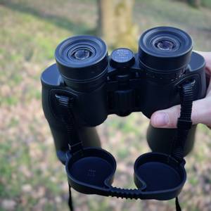 Nahaufnahme des Nikon Aculon A211 12x50 Fernglases mit befestigten Schutzkappen an den Objektiven. Die Kappen hängen an elastischen Riemen und sind dadurch gesichert.