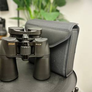 Das Nikon Aculon A211 12x50 Fernglas steht aufrecht neben seiner schwarzen Tragetasche. Die Tasche hat einen Klettverschluss zur sicheren Aufbewahrung.