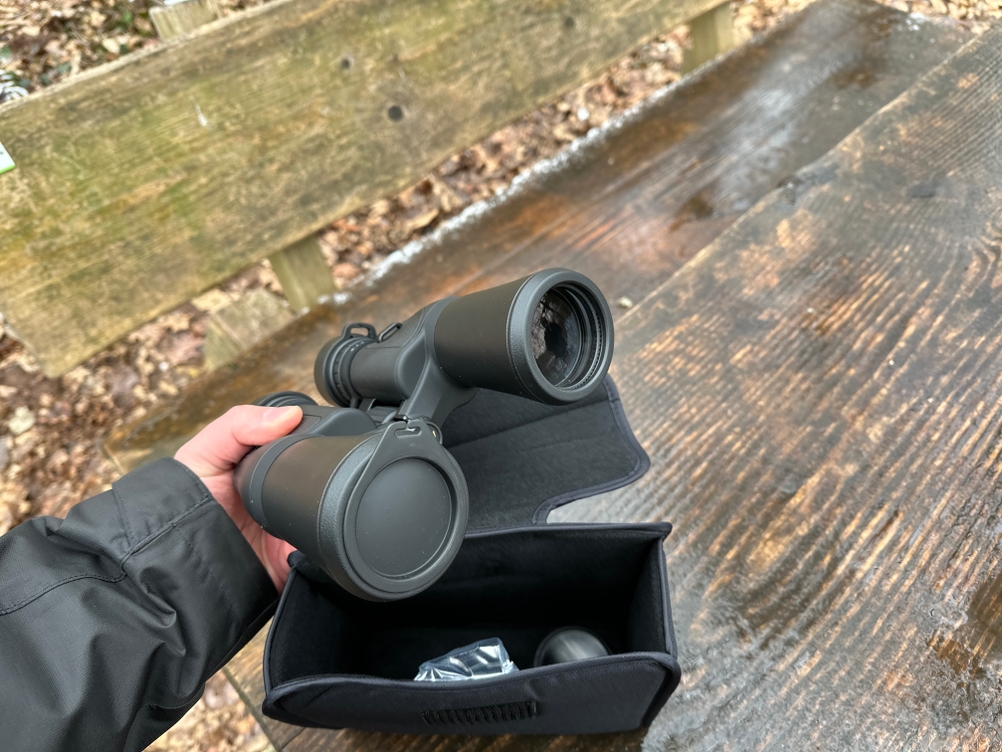 Eine Hand hält das Nikon-Aculon-A211-12x50-Fernglas. Eine Linse ist von einer Schutzkappe verdeckt. Im Hintergrund ein feuchter Holztisch und eine schwarze Tasche.