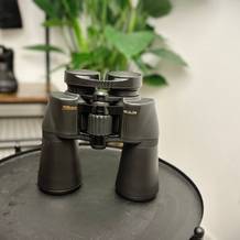 Das Nikon Aculon A211 12x50 steht auf einem Tisch, die Gummiarmierung und die Einstellräder sind gut sichtbar. Im Hintergrund sind Wanderschuhe und eine Pflanze zu sehen.