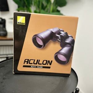 Die Verpackung des Nikon Aculon A211 12x50 zeigt eine Abbildung des Fernglases auf einem gold-braunen Hintergrund. Oben links ist das Nikon-Logo sichtbar.