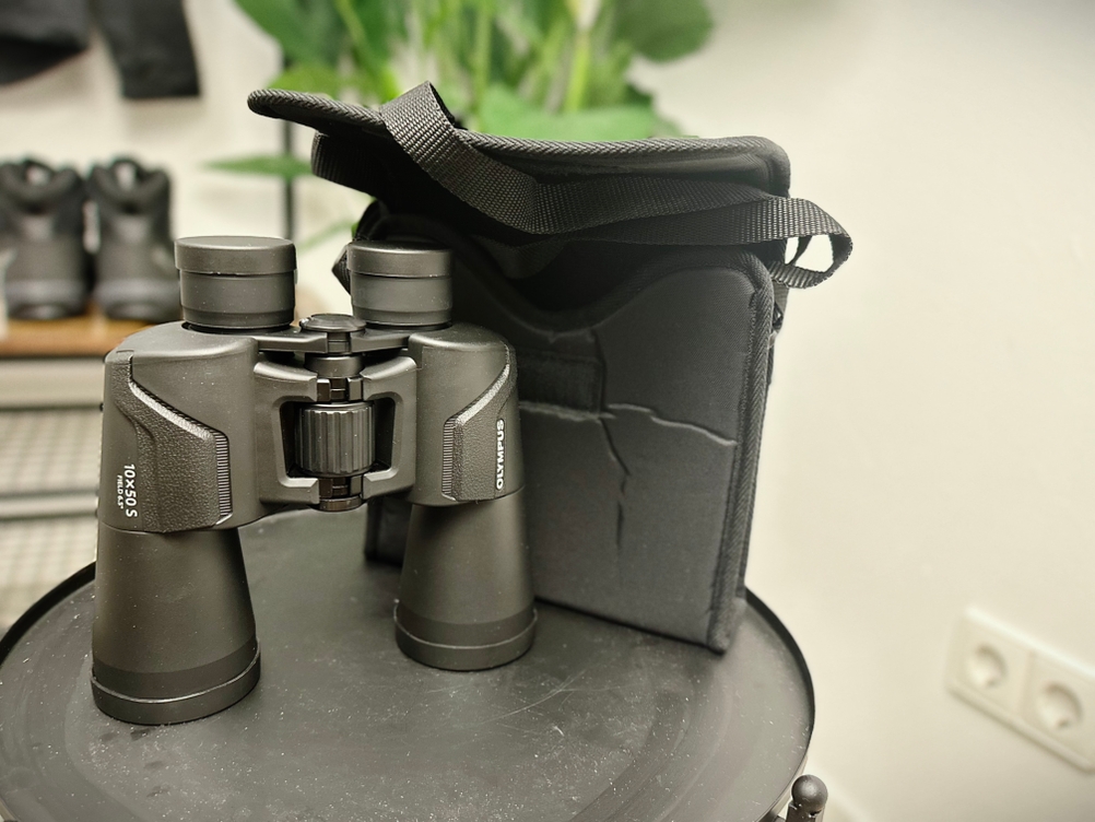 Das Olympus-10x50-S-Fernglas steht neben seiner mitgelieferten Tragetasche auf einem schwarzen Tisch. Beide sind für den Transport gedacht.