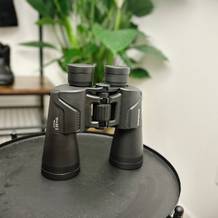 Das Olympus 10x50 S Fernglas steht auf einem Tisch, die Gummiarmierung und die Einstellräder sind gut sichtbar. Im Hintergrund sind Wanderschuhe und eine Pflanze zu sehen.