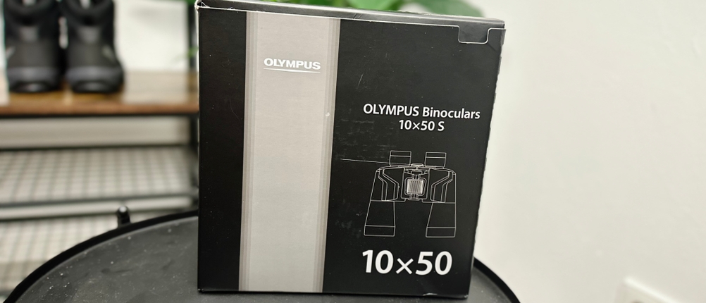 Die Verpackung des Olympus-10x50-S-Fernglases steht auf einem schwarzen Tisch. Im Hintergrund sind Wanderschuhe und eine Pflanze zu sehen.