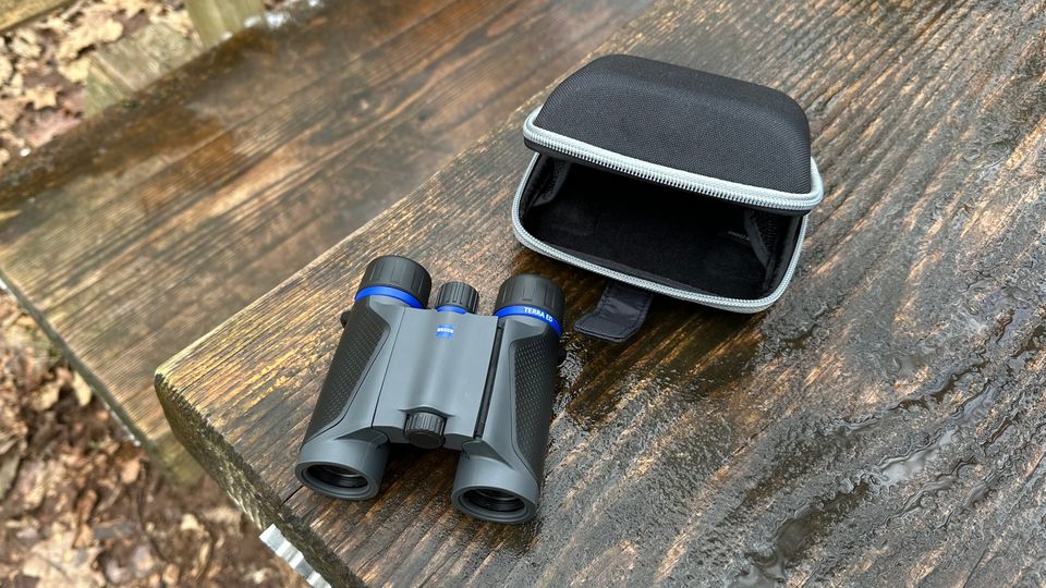 Das Zeiss Terra ED Pocket Fernglas liegt neben seiner kompakten Schutztasche auf einer nassen Holzbank im Freien. Die Tasche ist geschlossen und mit einem Reißverschluss gesichert.