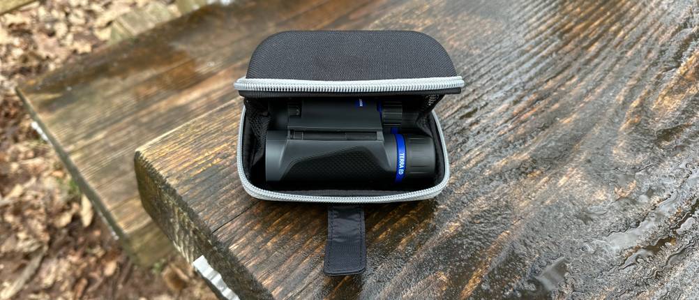 Das Zeiss-Terra-ED-Pocket-Fernglas liegt in seiner geöffneten Transporttasche auf einer Holzbank. Die gepolsterte Hülle schützt das Fernglas vor Stößen und Kratzern.