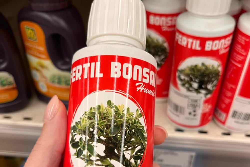 Hand hält vor Verkaufsregal eine rot bedruckte Flasche mit Fertil-Bonsai-Dünger im Test.