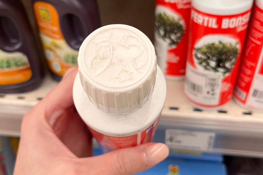 Hand hält vor einem Verkaufsregal eine Flasche mit Sicherheitsverschluss eines getesteten Bonsai-Düngers von Fertil in die Kamera.