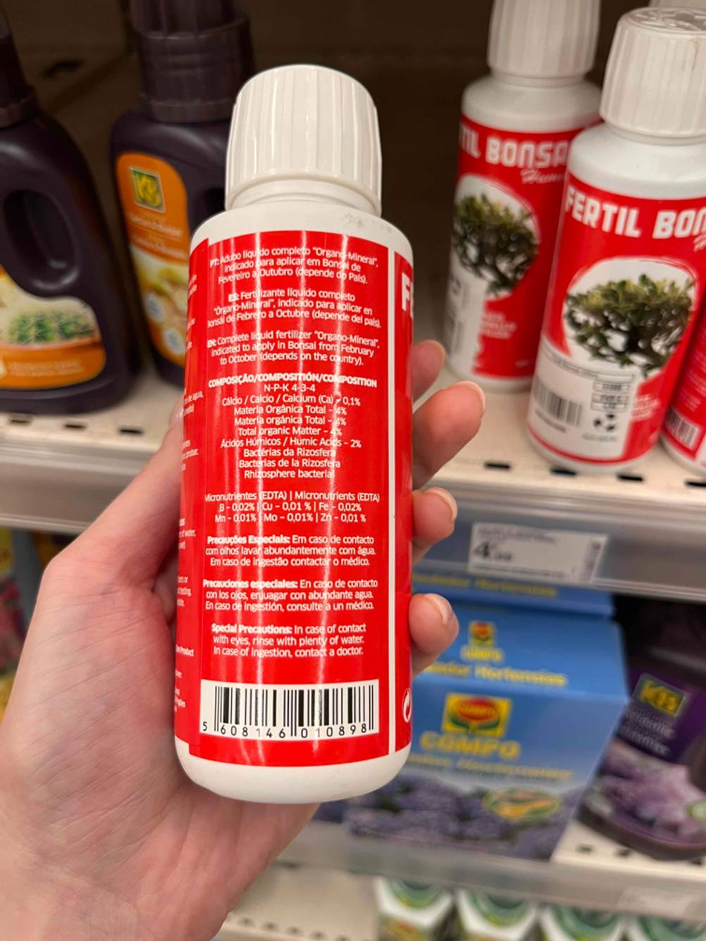 Bonsai-Dünger im Test: Hand hält rot Dünger-Flasche von Fertil mit Fokus auf Informationen zum Inhalt und zur Anwendung vor einem Verkaufsregal.