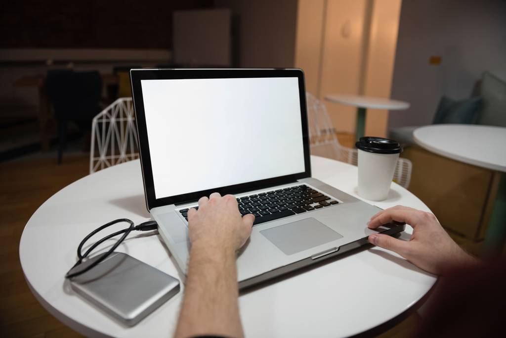 Festplattengehäuse im Test: Eine Person sitzt im Café an einem runden Tisch, auf dem ein Laptop, ein Festplattengehäuse aus Metall und ein Kaffeebecher positioniert sind.