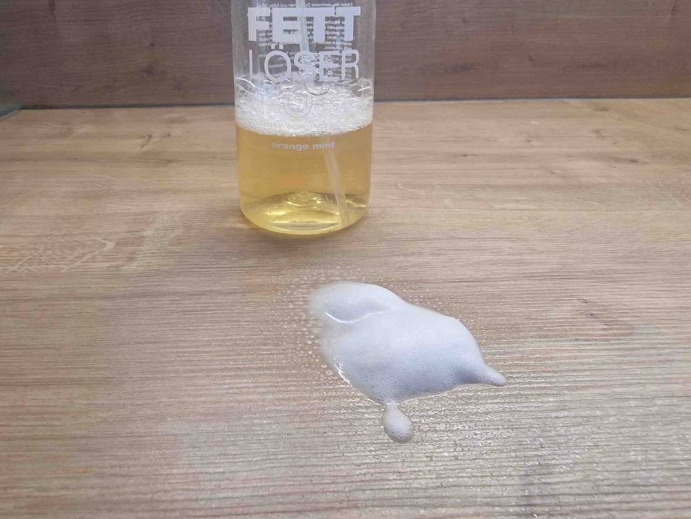 Getestet: Etwas schäumender Fettlöser wurde auf einen Holztisch gesprüht, auf dem im Hintergrund die Flasche steht.