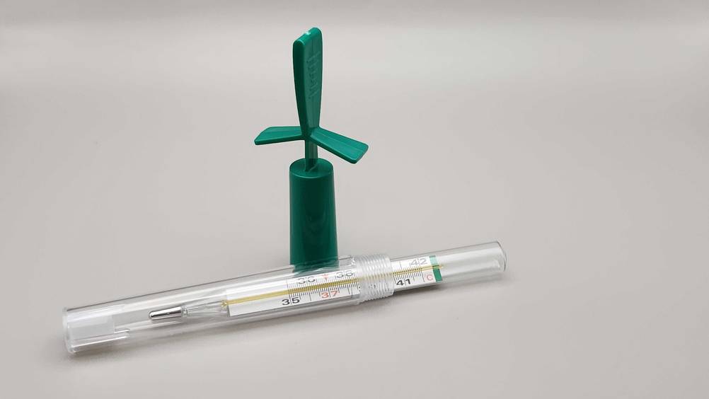 Ein getestetes Fieberthermometer analog in seiner Schutzhülle neben der Verschlusskappe auf einer weißen Fläche.