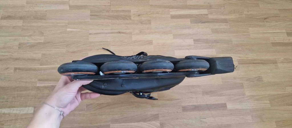 Eine Damenhand hält einen schwarzen getesteten Inline-Skate für Damen, mit Fokus auf die Unterseite der Rollen, über einem Holzboden.