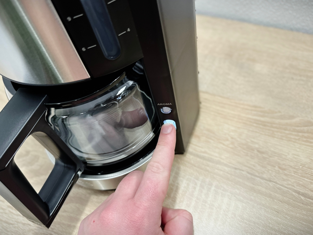 Eine Hand drückt den Einschaltknopf der Krups KM3210 Filterkaffeemaschine. Die Tasse ist bereitgestellt, die Maschine startet den Brühvorgang.