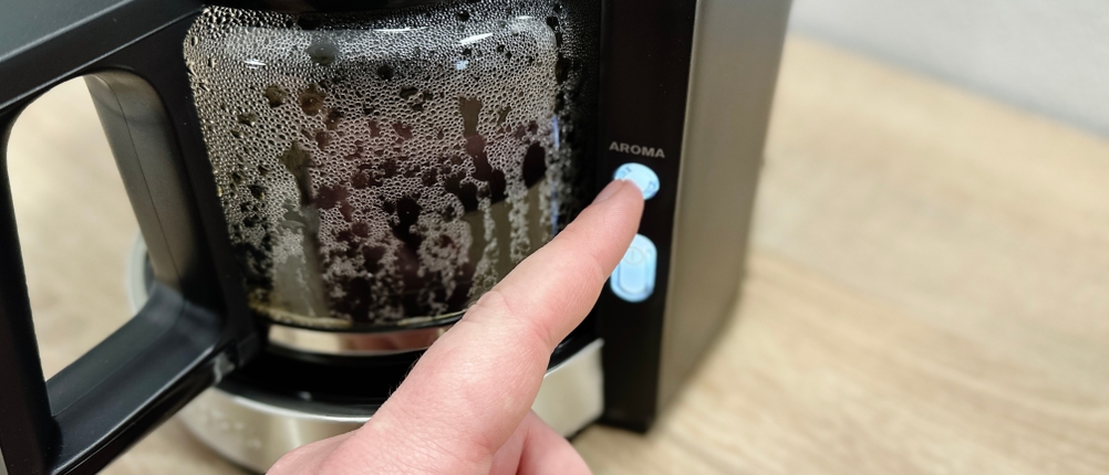 Eine Hand aktiviert die Aroma-Funktion der Krups KM3210 während des Brühvorgangs. Die Glaskanne ist bereits teilweise gefüllt.