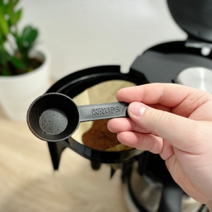 Ein Kaffeelot der Marke Krups wird über einem Papierfilter mit Kaffeepulver gehalten. Im Hintergrund ist der offene Filtereinsatz der Maschine zu sehen.