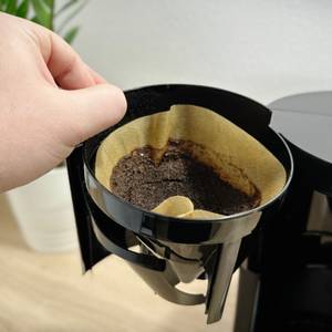 Ein gebrauchter Papierfilter mit feuchtem Kaffeepulver wird aus der Krups KM3210 entnommen. Die Maschine ist geöffnet.