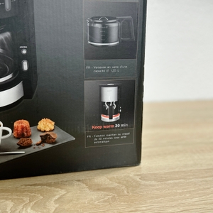 Detailausschnitt der Verpackung der Krups KM3210 mit Informationen zu Glaskanne und Warmhaltefunktion. Darunter ist eine Tasse Kaffee mit Gebäck zu sehen.