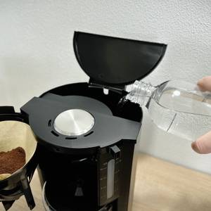 Eine Hand füllt Wasser aus einer Flasche in den offenen Wassertank der Krups KM3210 ein. Daneben befindet sich ein Filtereinsatz mit Kaffeepulver.