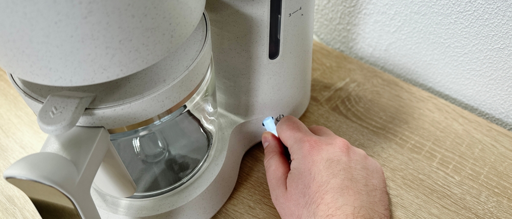 Eine Hand schaltet die Philips-HD5120-Filterkaffeemaschine mit einer seitlichen Taste ein. Die Maschine steht auf einer Holzplatte.
