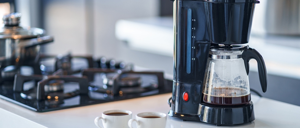 Zu sehen ist eine Küchenzeile, auf der eine Filterkaffeemaschine steht sowie zwei mit Kaffee gefüllte Tassen. Im Hintergrund ist ein Gasherd zu sehen.