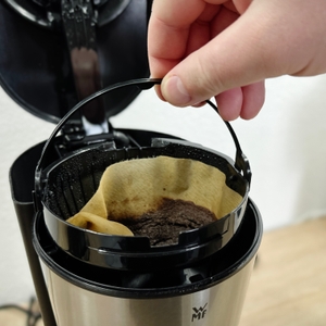 Ein gebrauchter Papierfilter mit nassem Kaffeesatz wird aus der WMF Bueno entnommen. Der Filterhalter ist schwarz und rund.