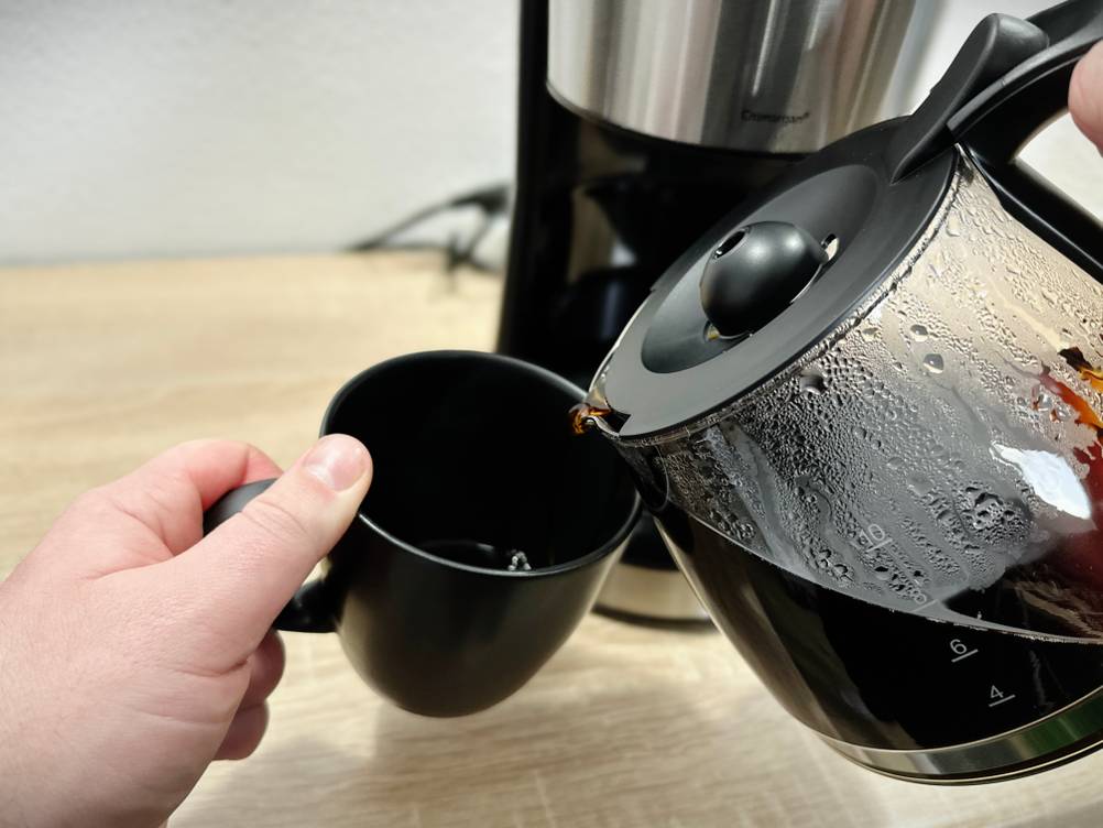 Kaffee wird aus der WMF-Bueno-Glaskanne in eine schwarze Tasse gegossen. Der Kaffee läuft direkt und mit leichtem Tropfen.
