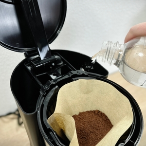 Wasser wird aus einer Flasche in den geöffneten Wassertank der WMF Bueno gegossen. Der Papierfilter mit Kaffeepulver ist bereits eingelegt.