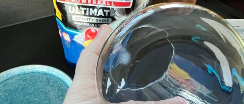 Eine Hand hält ein Glas mit leichter Verschmutzung fest. Im Hintergrund die Verpackung der Finish-Ultimate-Infinity-Shine-Spülmaschinentabs.