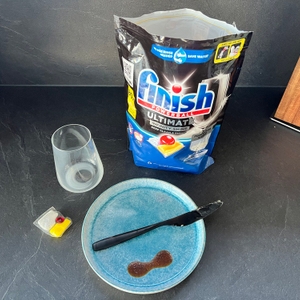 Auf einer Küchenarbeitsplatte stehen ein Teller mit einem eingetrocknetem Fleck Ketchup, ein Messer mit Butter, ein Glas mit eingetrockneter Milch, ein Finish Ultimate Infinity Shine Spülmaschinentab und die Packung der Spülmaschinentabs