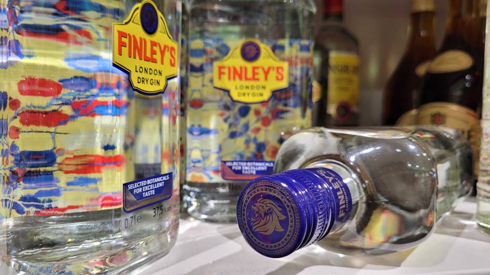 Gin-Test: Finleys London Dry Gin in einer transparenten flachen Flasche mit lila Schraubverschluss liegt vor stehenden Flaschen.