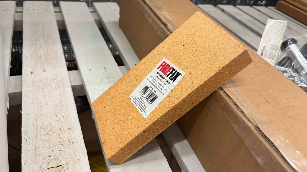 Auf einer weißen Palette im Baumarkt lehnt der getestete Schamottstein in Hellgelb an einem braunen Karton.