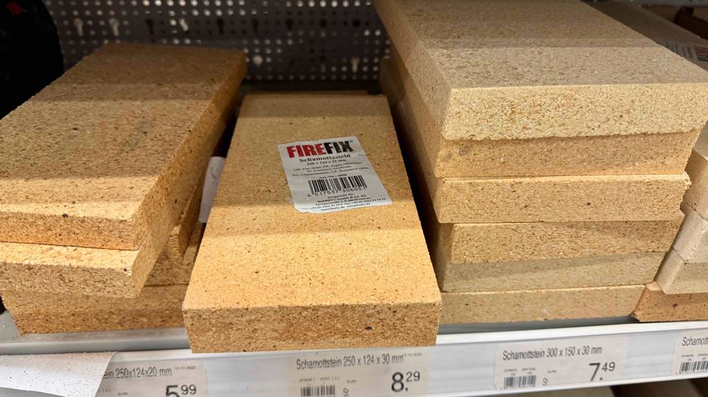 Mehrere Schamottsteine im Test liegen neben- und übereinander in einem Verkaufsregal, bestückt mit unterschiedlichen Preisschildern. 