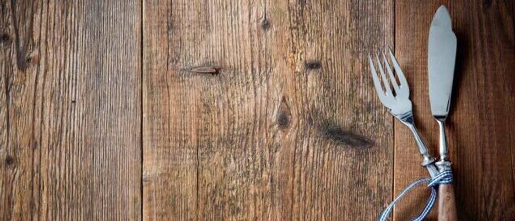 Fischbesteck-Test: Auf einer dunkelbraunen Holzplatte liegt ein Fischbesteck überkreuz sowie ein Anhänger mit zwei Fischen aus Metall.
