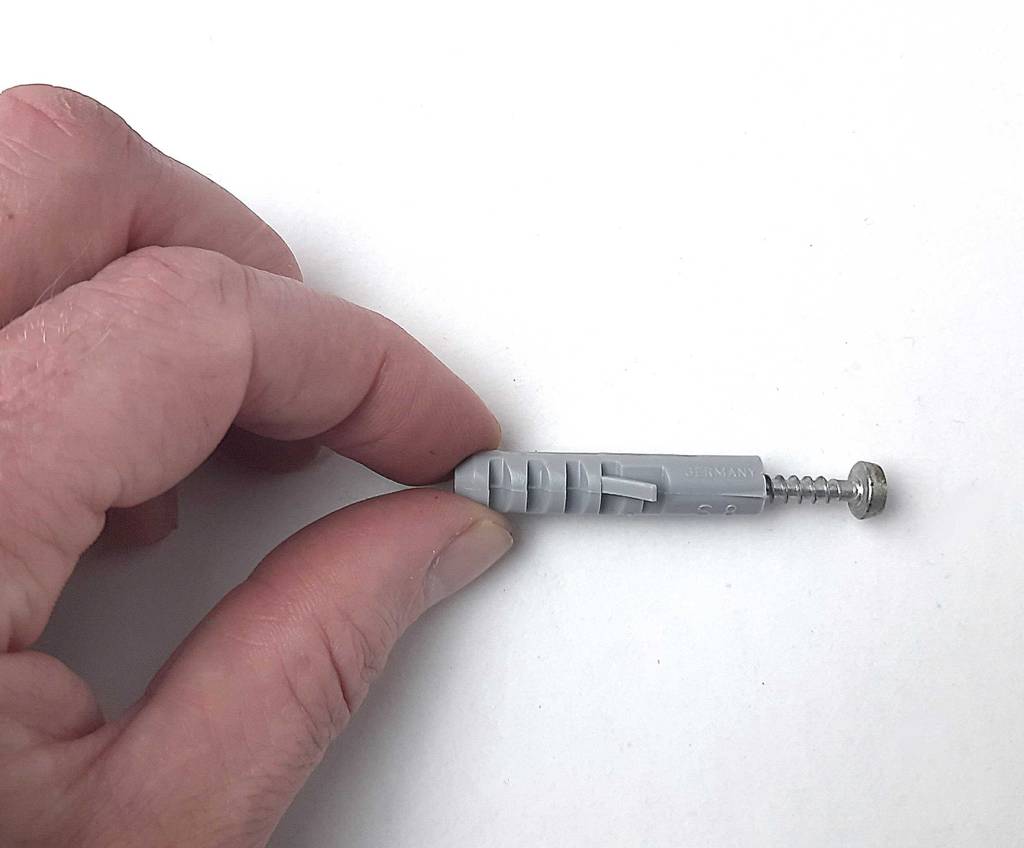 Zwischen zwei Fingerspitzen befindet sich der getestete Dübel in Grau inklusive eingeschobener Schraube.