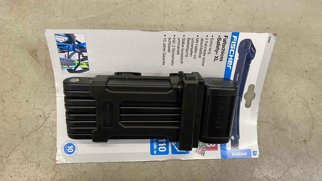 Faltschloss im Test: Das Faltschloss Safety XL (110 cm Länge) von Fischer inklusive Schlüssel liegt auf einer grauen Oberfläche.