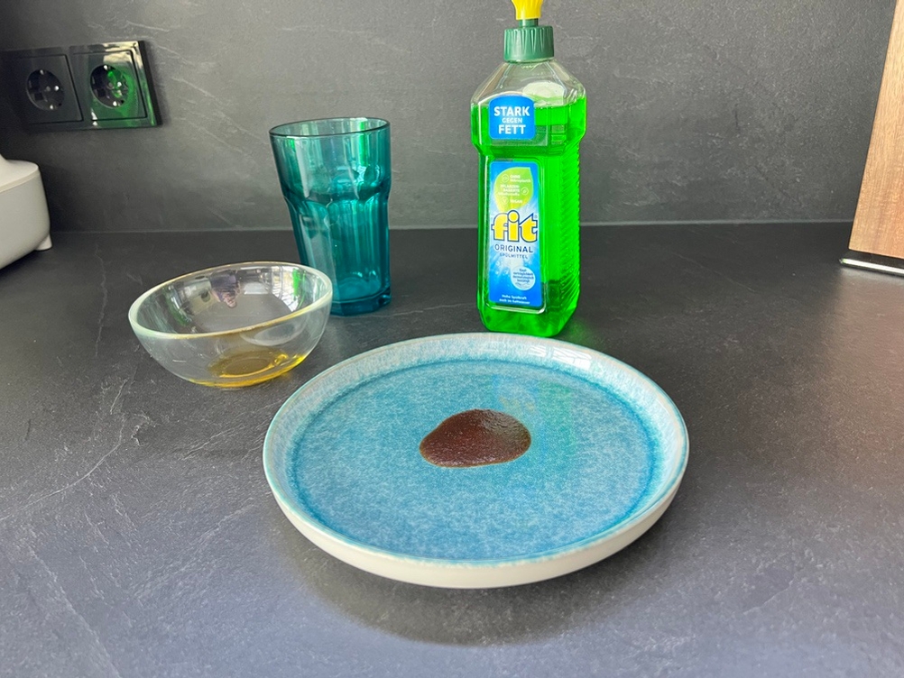 Die Flasche Fit-Spülmittel Original steht neben einem Teller mit eingetrockneten Ketchupresten, einem Glas mit eingetrockneter Milch und einer Schale Olivenöl auf einer Küchenarbeitsplatte.