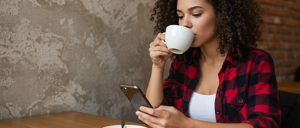 flanellhemd-test: Frau im schwarz-rot karierten Flanellhemd sitzt im Café und trinkt Kaffee, während sie auf ihr Handy schaut.
