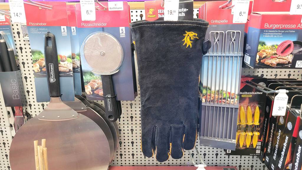 Ein Paar Grillhandschuhe hängt zum testen in einem Lochwandregal zwischen anderen Grillutensilien.