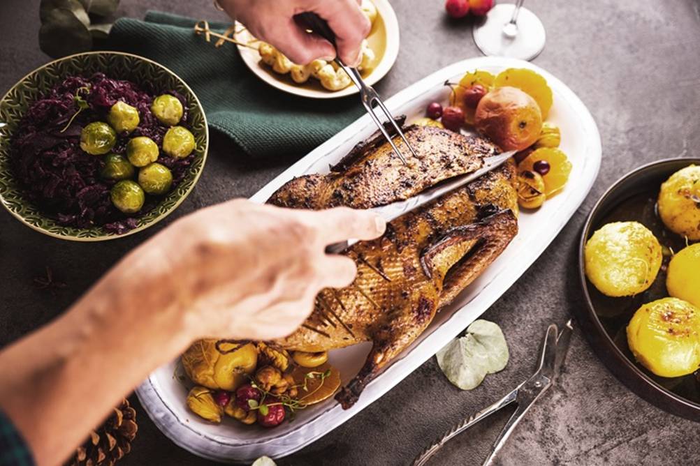 Ein Mann schneidet einen Braten mit Hilfe einer Fleischgabel und eines Tranchiermessers. Auf dem Tisch steht außerdem noch verschiedenes Gemüse.