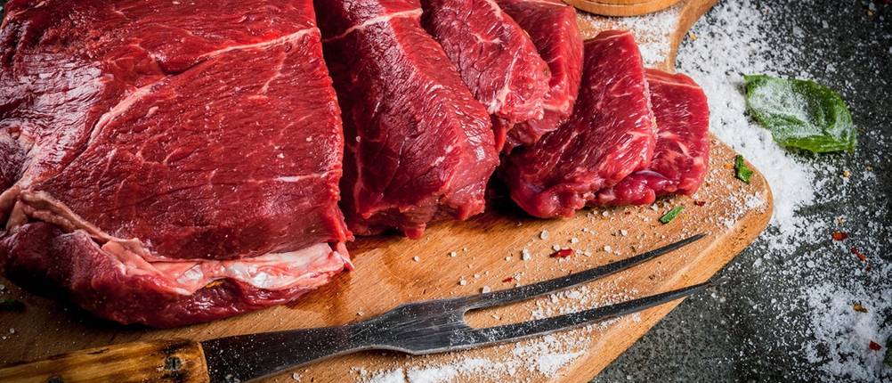 Eine Fleischgabel mit Holzgriff liegt auf einem Holzbrett neben rohem, zur Hälfte geschnittenem Fleisch.