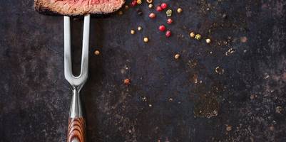 Zwei Stück Fleisch sind auf einer Fleischgabel und liegen auf einem schwarzen Untergrund mit Pfeffer neben der Fleischgabel.