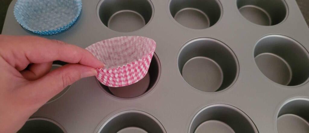 Muffinformen im Test: Hand legt eine rosafarbene Muffinform aus Backpapier in eine anthrazitfarbene Aluminiumform, die zwölf Mulden aufweist.