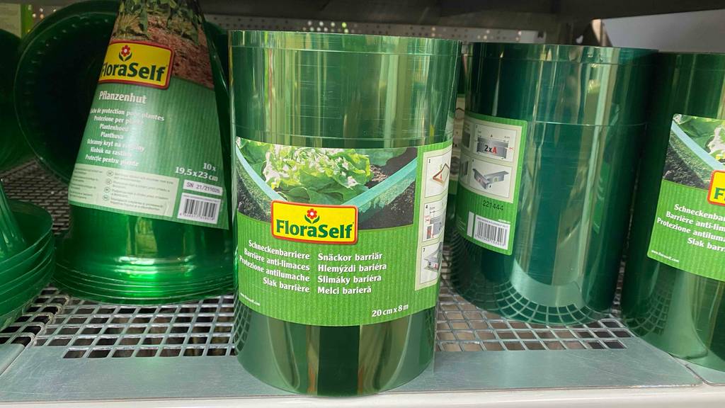 Schneckenzaun-Test: Mehrere Verpackungen mit eingerollten Schneckenzäunen in acht Meter Länge und zwanzig Zentimeter Höhe stehen nebeneinander in einem Verkaufsregal neben Pflanzenhüten.
