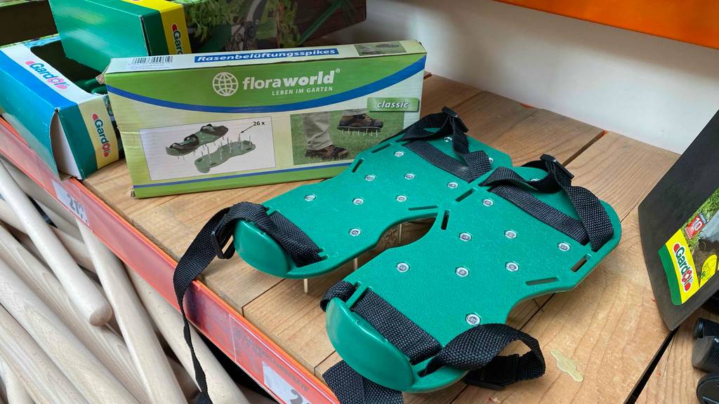Im Test: Rasenlüfter-Schuhe in Grün mit schwarzen Gurten stehen neben weiteren Gartenprodukten auf einem Verkaufsregal.