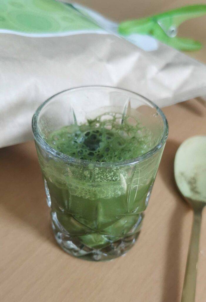 Gerstengras-Pulver im Test: ein transparentes Trinkglas mit grüner Flüssigkeit steht neben einem Teelöffel, der auf einem Holztisch liegt.