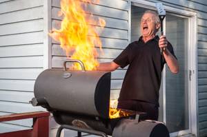 Ein Mann steht mit aufgerissenen Augen und offenem Mund und einem Pfannenwender in der Hand vor einem Grill, aus dem eine hohe Flamme sticht. Im Hintergrund ist ein Haus sowie eine Terrassentür zu sehen.