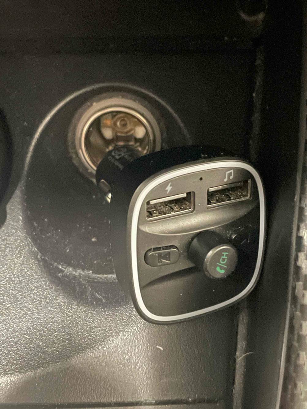Ein schwarzer FM-Transmitter inklusive Adapter vor einem Zigarettenanzünder-Eingang.