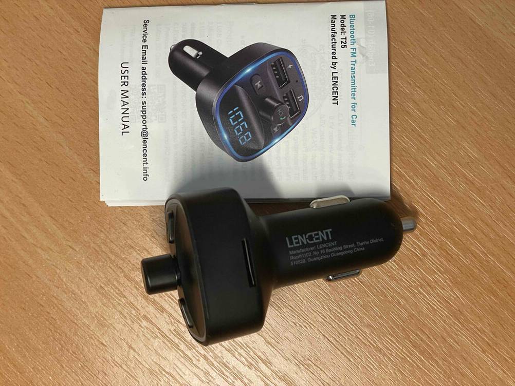 Ein schwarzer, getesteter FM-Transmitter liegt neben seinem Handbuch auf einer Holzfläche.